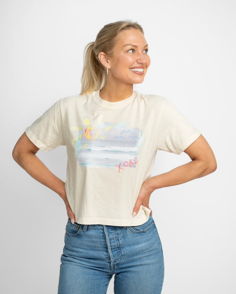 Happy blonde woman wearing a cream colored t shirt tucked into her blue jeans. T shirt has a beach scene printed on the front of it with the letters XOXO in pink.