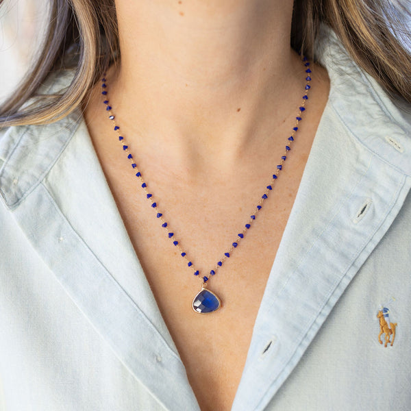 Model wearing necklace made of tiny blue beads with a large, teardrop, blue stone pendant