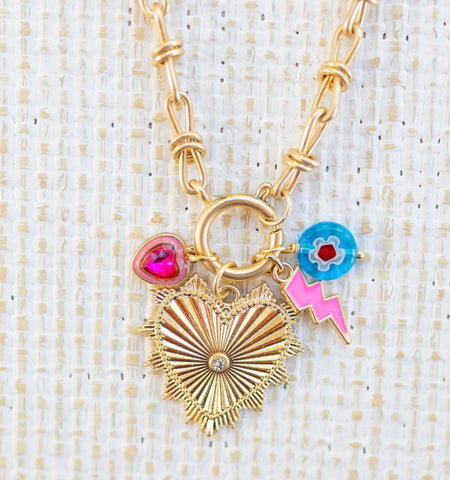 Picture of gold toggle chain with a large gold heart pendant with a diamond in the center. Three charms featured are a blue bead with a white and red flower, a pink lightening bold and pink heart stone charm.