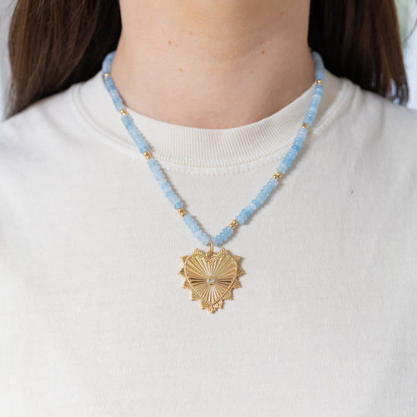 Picture of model wearing a light blue colored beaded necklace with gold accent beads. Necklace has large gold heart pendant with diamond stud at center.