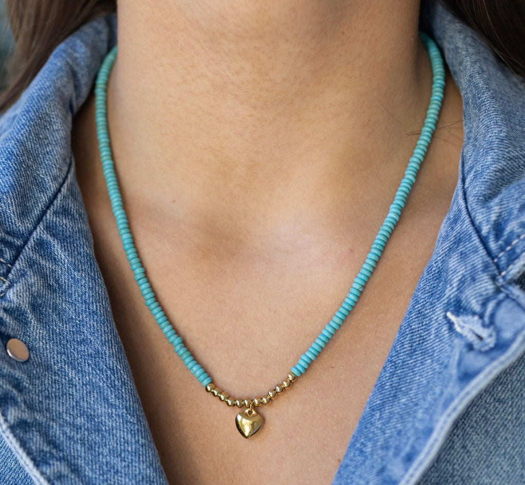 Model wearing blue beaded necklace with small gold heart pendant