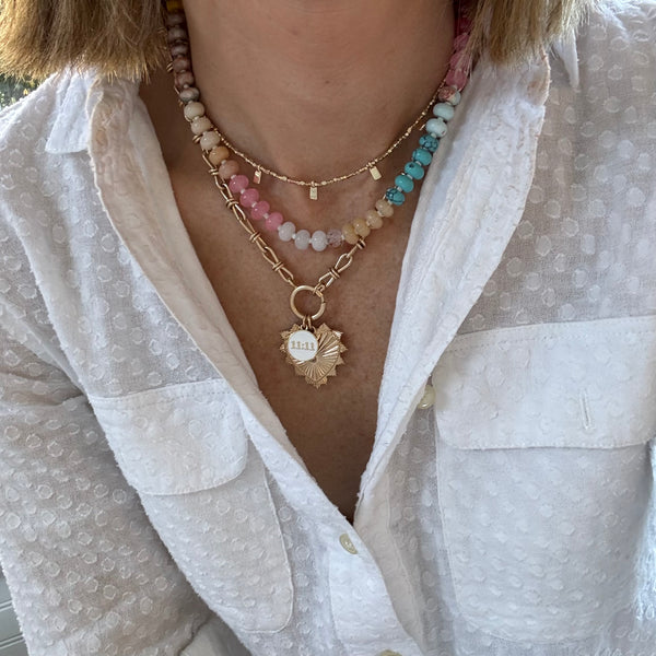 Picture of model's neck wearing a necklace stack. First is a delicate gold necklace with gold tab beads, second muted tan, rose, white and aqua colored bead necklace and the last is a chunky link necklace with a large gold heart pendant and a smaller white pendant reading "11:11"