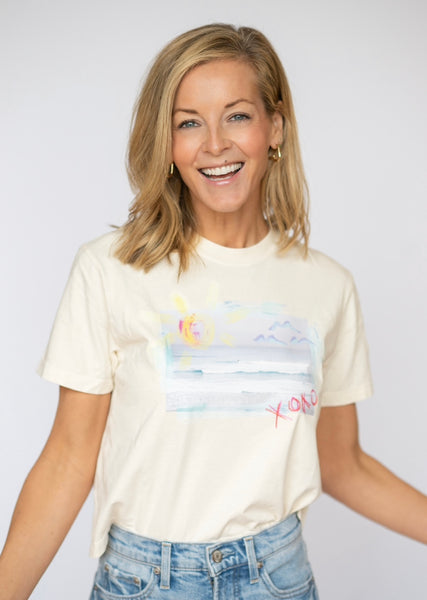 Happy blonde woman wearing a cream colored t shirt tucked into her blue jeans. T shirt has a beach scene printed on the front of it with the letters XOXO in pink.