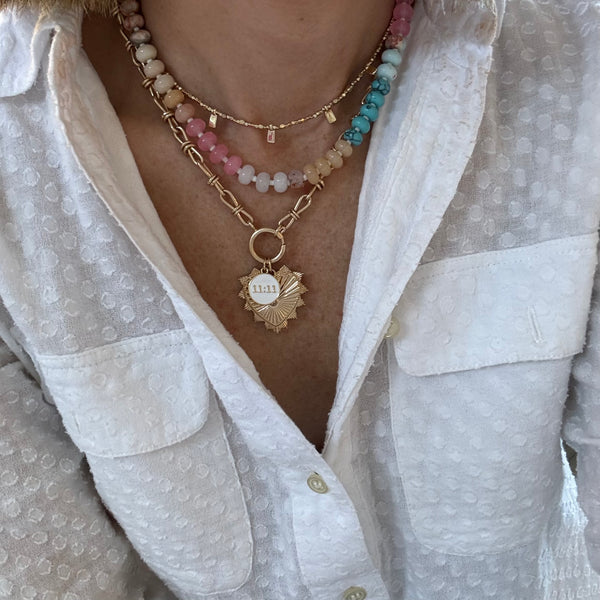 Picture of model's neck wearing a necklace stack. First is a delicate gold necklace with gold tab beads, second muted tan, rose, white and aqua colored bead necklace and the last is a chunky link necklace with a large gold heart pendant and a smaller white pendant reading "11:11"