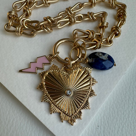 Picture of gold chain link charm necklace.  Necklace has a large gold heart pendant with a diamond at its center. Two smaller charms include a dark blue bead and a pink lightening bolt.