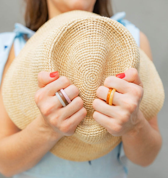 Woman holding a straw sun hat in her arms against her chest and wearing orange, blue, and pink rings 