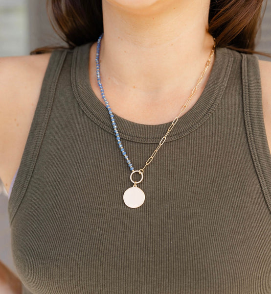 Woman wearing a necklace with gold links as one half of the chain and the other half tiny blue beads with a circular gold medallion in the middle