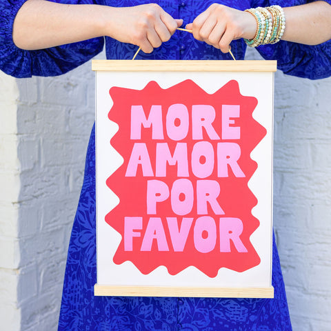 Girl against a wall wearing a blue dress holding a canvas hanging art piece with the text "more amor por favor"