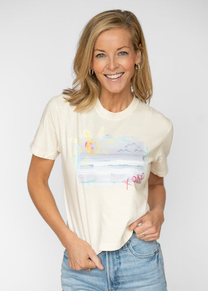 Happy blonde woman wearing a cream colored t shirt tucked into her blue jeans. T shirt has a beach scene printed on the front of it with the letters XOXO in pink.