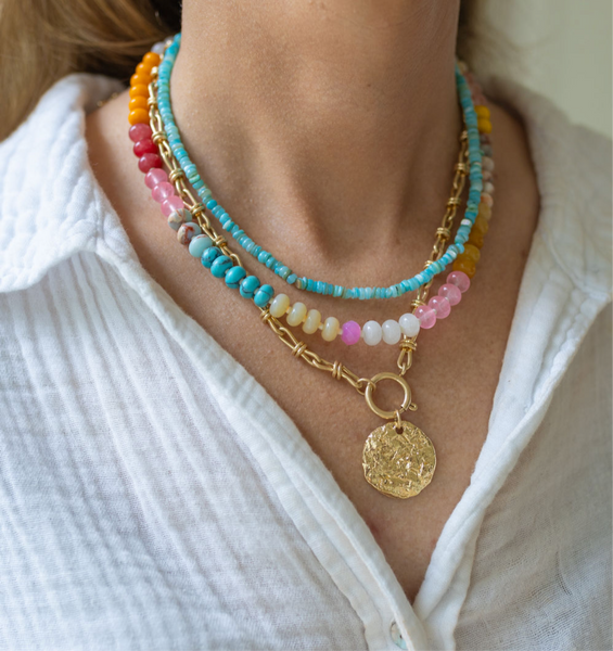 Woman wearing three necklaces: a gold chunky chained necklace sitting on a table with a large gold medallion attached to it, a multi-colored stone beaded short necklace, and a small blue beaded choker