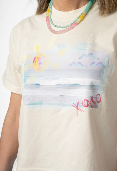 Picture of a woman wearing a cream colored Tshirt with a beach scene printed on it. It also has the letters XOXO written on it. She is wearing a semi-precious stone beaded necklace in light pink, yellow and teal, layered with a tiny teal beaded necklace.