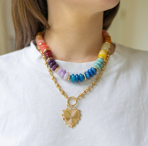 Model wearing large multicolored beaded necklace stacked with front clasping gold chain necklace with a heart pendant