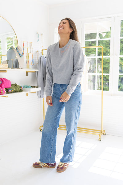 Woman standing in a house posing wearing a gray sweatshirt with blue stitching that says  "Joie De Vivre"