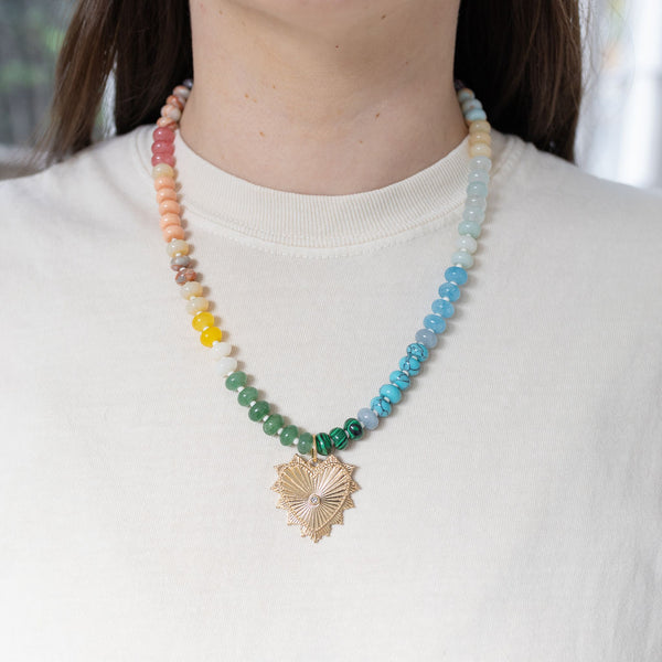 Model wearing a multi-colored beaded necklace of pink, peach, yellow, white, green and blues. Necklace has a large gold heart pendant necklace with a diamond at its center.