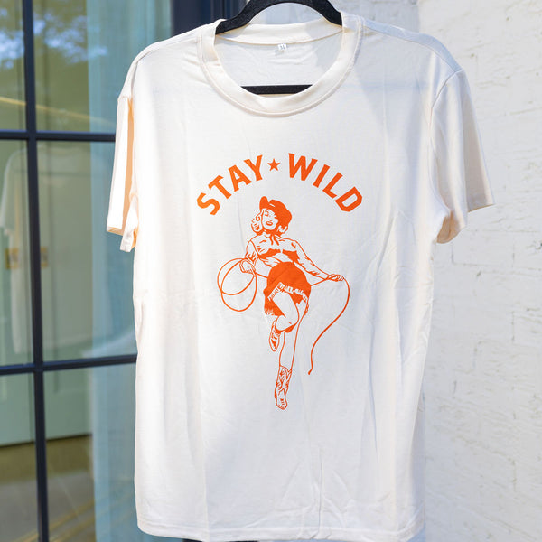 A cream colored t-shirt with a graphic of an orange cowgirl with a cowboy hat and lasso with the text "stay wild" hanging on a black coat hanger 