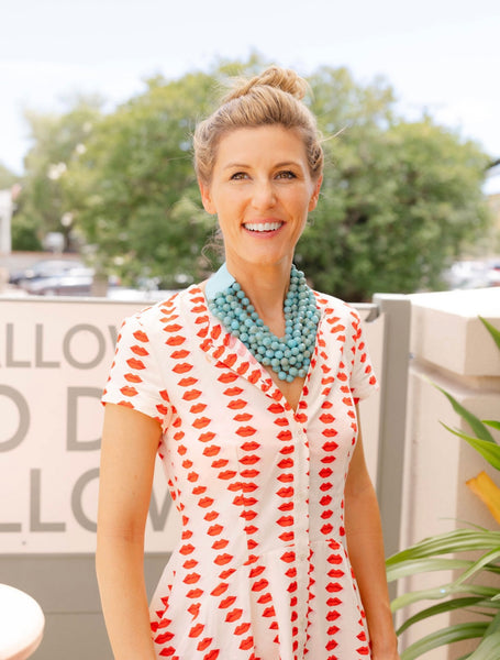 Woman outside smiling wearing a white dress with a red lips print and a big blue stone layered necklace around her neck