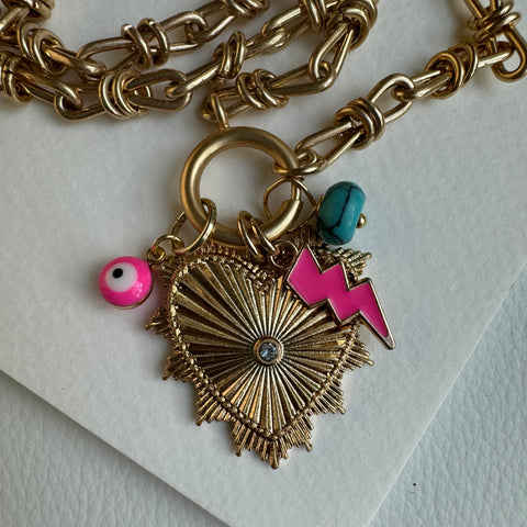 Picture of gold chain link charm necklace.  Necklace has a large gold heart pendant with a diamond at its center. Three smaller charms include a turquoise bead, a pink "evil eye" and a pink lightening bold.