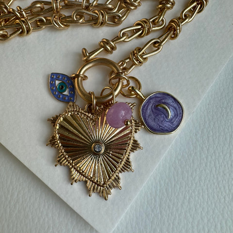 Picture of gold chain link charm necklace.  Necklace has a large gold heart pendant with a diamond at its center. Three smaller charms are also attached. One a small pink bead, another a blue shimmery disc with a silver crescent at its center and the last a blue "evil eye".