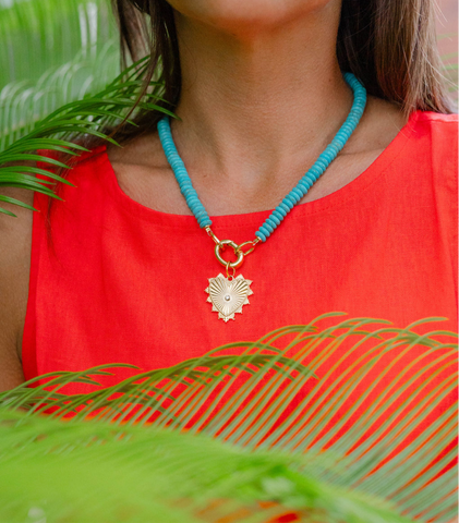 Woman wearing a big blue beaded necklace with a gold heart shape medallion attached to it