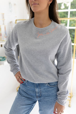 Woman standing in a house posing wearing a gray sweatshirt with blue stitching that says  "Joie De Vivre"