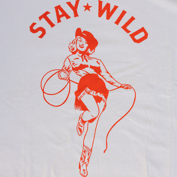 A cream colored t-shirt with a graphic of an orange cowgirl with a cowboy hat and lasso with the text "stay wild"