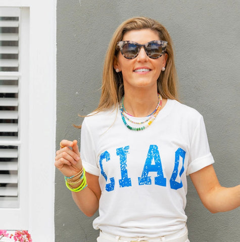 Woman looking away wearing a white shirt with blue print text that says "Ciao" wearing beaded colorful bracelets and necklaces