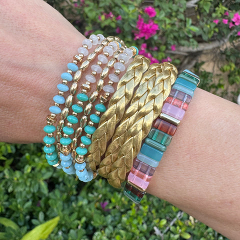 Picture of wrist wearing various bracelets. 3 rose and turquoise, 2 gold beaded, 3 gold woven and 1 large rectangle beaded bracelet.