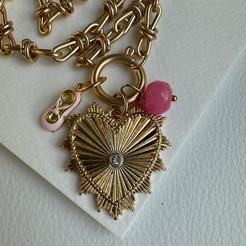 Picture of gold chain link charm necklace.  Necklace has a large gold heart pendant with a diamond at its center. Two smaller charms include a cut pink rose colored bead and an oval light pink and gold charm that resembles a ballerina slipper