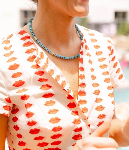 Woman outside wearing a white dress with a red lips print and a blue/gold beaded necklace around her neck