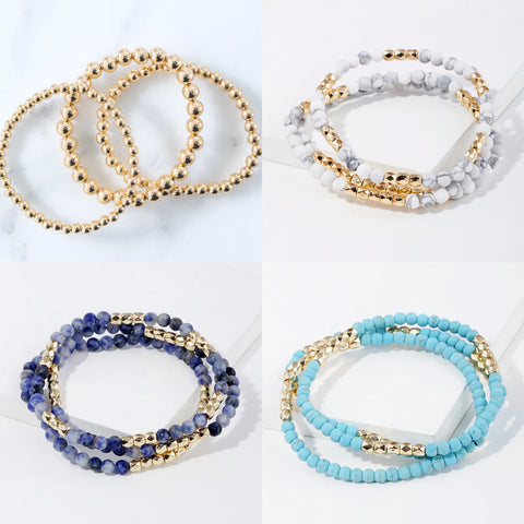 Picture of various beaded bracelet sets. Each set has three bracelets. One set is in all gold, one in white marble and gold, one in ark blue marble and gold and the last in light blue marble beads and gold.