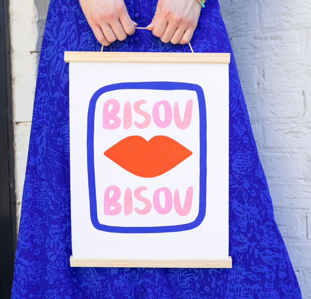 Girl against a wall in a blue dress holding a canvas sign with the words "Bisou Bisou" with red lips in a blue line rectangle 