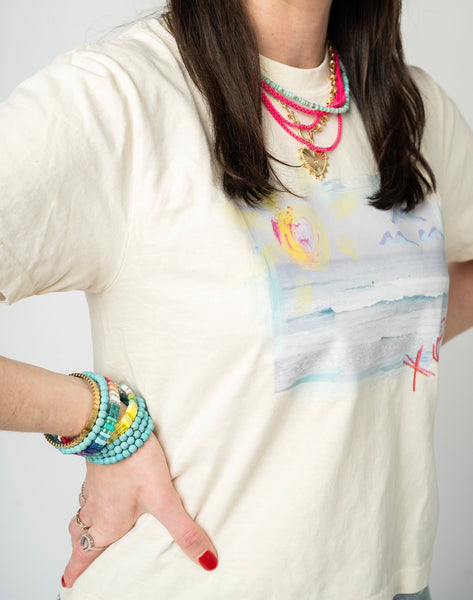 Picture of a woman wearing a cream colored t shirt with a picture of a beach scene. She also is wearing a layered stack of beaded necklaces in bright coral, light blue and a gold toggle heart necklace. She is also wearing a beaded turquoise and yellow bracelet bundle.