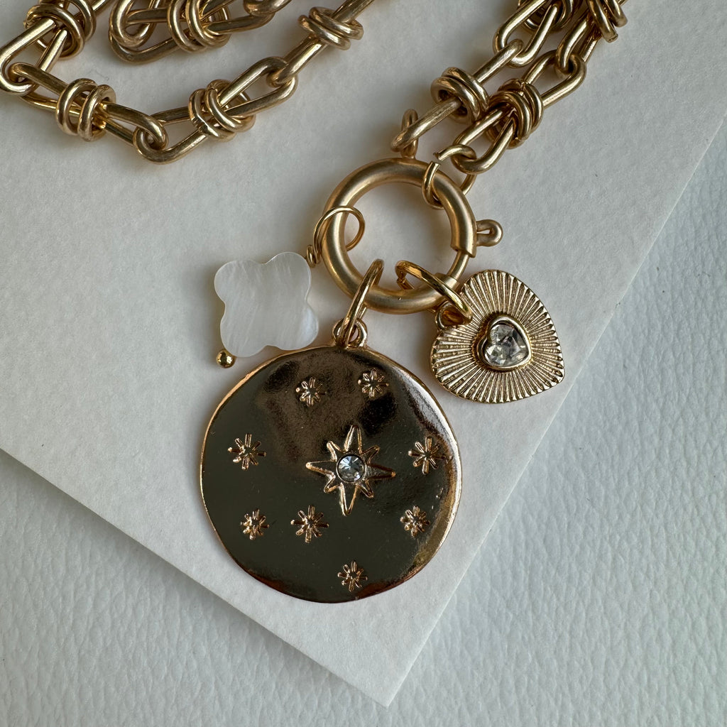 Picture of gold chain link charm necklace.  Necklace has a large gold disc with stars engraved and a diamond in the center of the largest star.  Two smaller charms include a white four petal clover charm and a small textured gold heart with a diamond heart at its center.