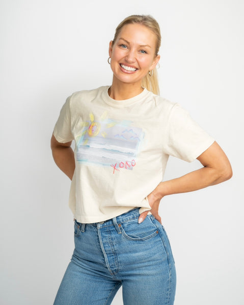 Happy blonde woman wearing a cream colored t shirt. T shirt has a beach scene printed on the front of it with the letters XOXO in pink.