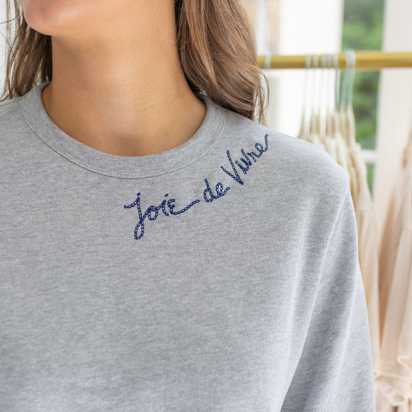 Woman wearing a gray sweatshirt with blue stitching that says  "Joie De Vivre"