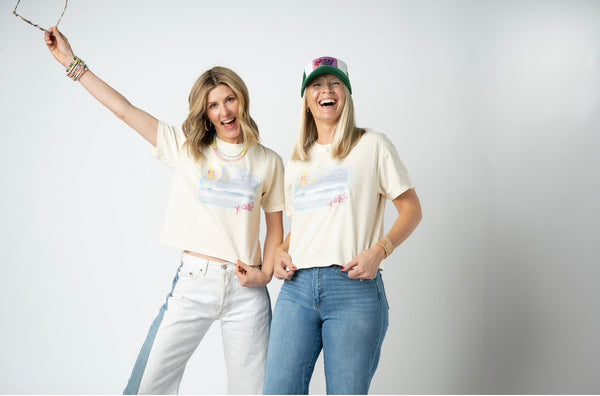 Picture of two women wearing jeans and matching beige cropped t shirts that have a beach scene. Women appear to be very happy and are accessorizing with trucker hats, sunglasses, statement jeans and beaded necklaces.