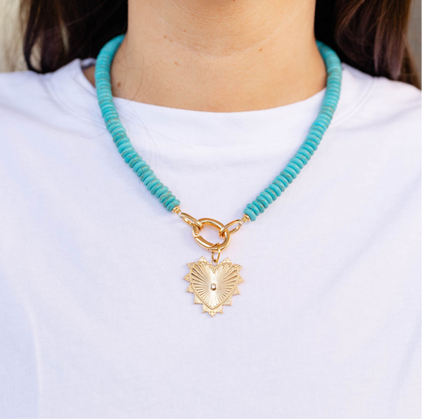 Woman wearing a big blue beaded necklace with a gold heart shape medallion attached to it