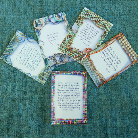 Five affirmation cards with necklaces/beads printed around the border laying on a green surface 
