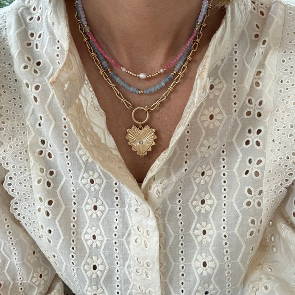 Picture of model wearing a necklace stack including a dainty blue and gold beaded necklace, a rose quartz and gold beaded necklace with a fresh water pearl center and a gold chain charm necklace with a large gold heart pendant with a diamond stud at its center.