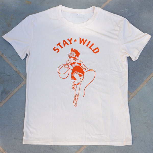 A cream colored t-shirt with a graphic of an orange cowgirl with a cowboy hat and lasso with the text "stay wild" laying on the ground