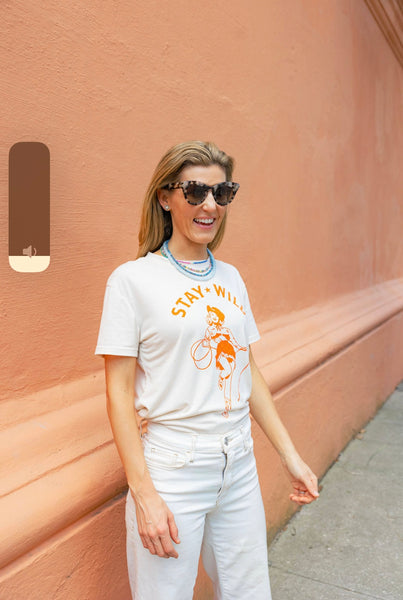 Woman wearing sunglasses and a cream shirt with an orange print of a cowgirl with a lasso and the text "Stay Wild" with multiple blue beaded necklaces