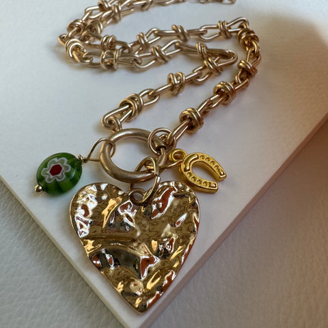 Picture of chunky gold chain necklace with a large gold heart pendant. Necklace also has two other charms attached, one a golden horseshoe and the other a green bead with a flower design.