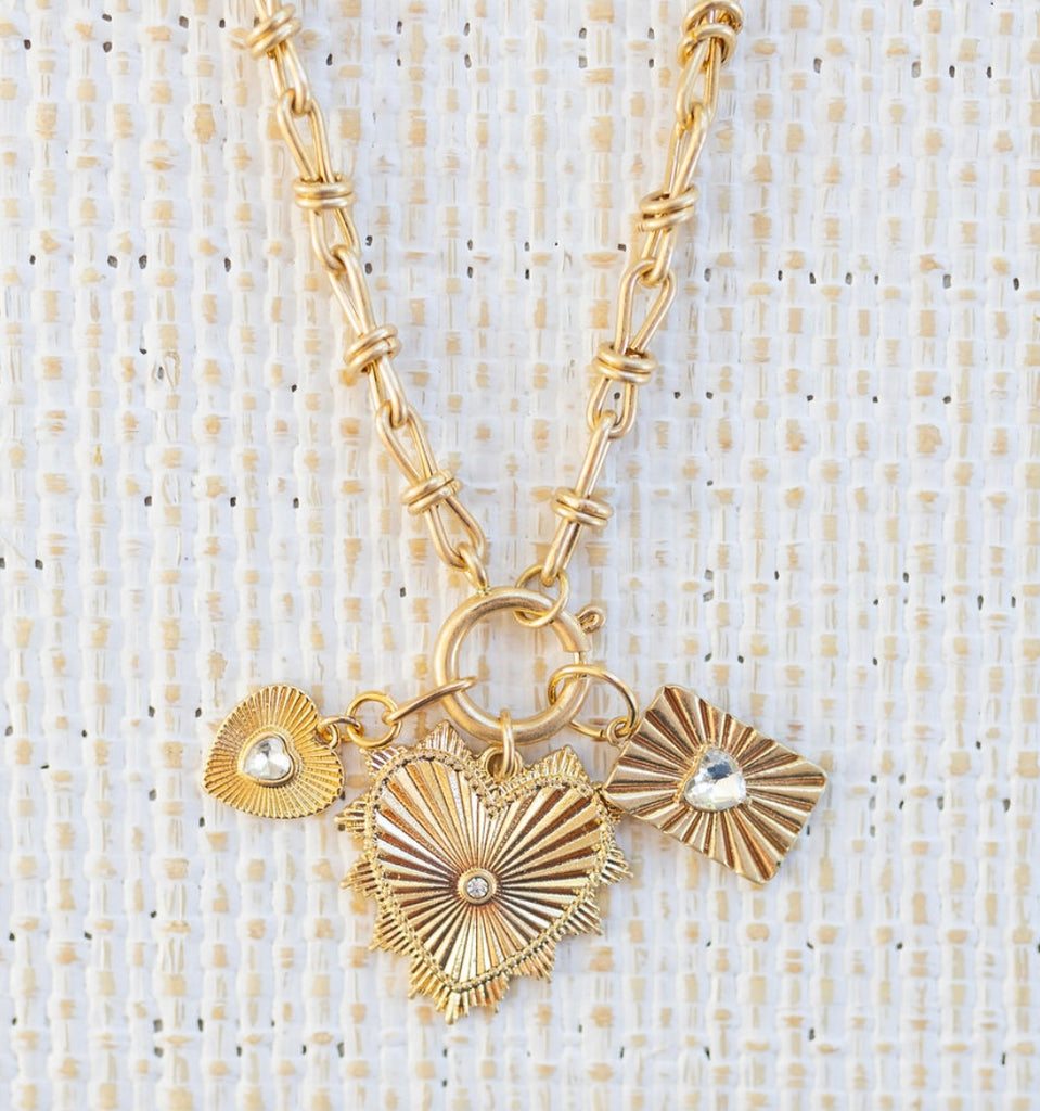 Picture of gold toggle chain with three pendants attached. One large gold heart with a diamond in the center, one small gold heart with a heart diamond in the center and one textured rectangle with a diamond heart in the center.