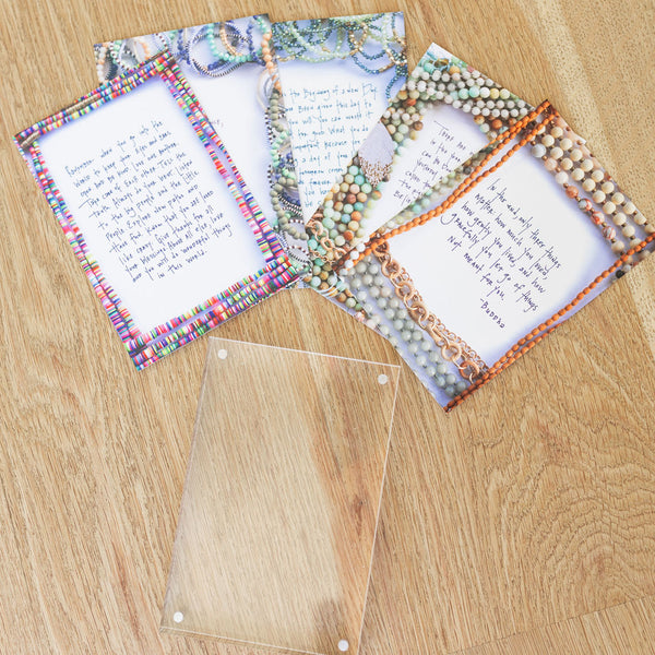 Five affirmation cards with necklaces/beads printed around the border with a clear card sleeve laying on a wooden surface 