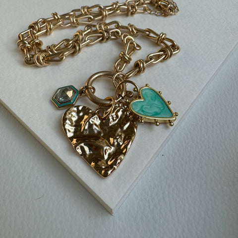 Picture of chain link gold charm necklace.  Necklace has a large textured gold heart pendant with two smaller charms near it. One of an turquoise heart with a studded gold border and a diamond charm with a turquoise and gold border.