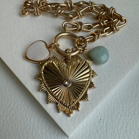 Picture of gold chain link charm necklace.  Necklace has a large gold heart pendant with a diamond at its center. Two smaller charms include a muted turquoise bead and a white heart charm with a gold border.