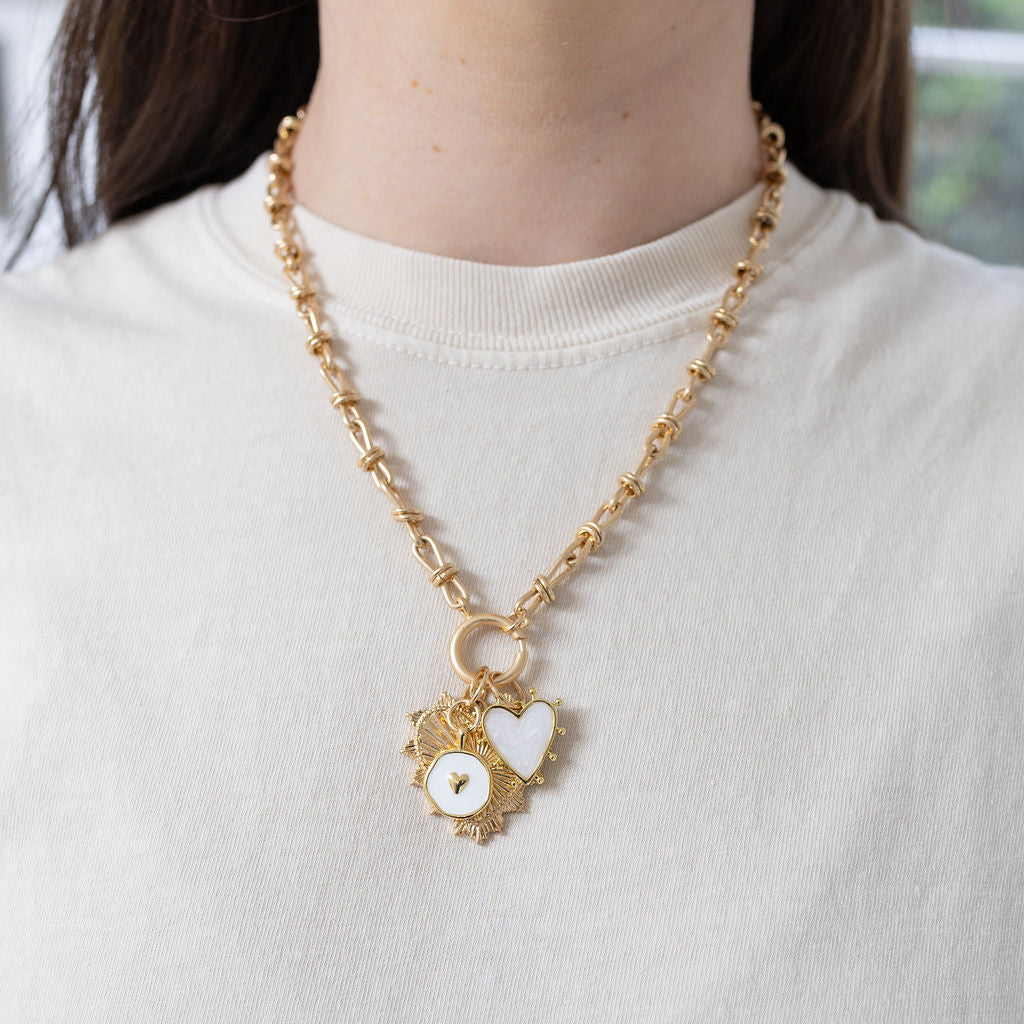 Model wearing a gold toggle chain with a large heart pendant with a diamond at the center Necklace has two more pendants, one a white heart and the other a white circle.