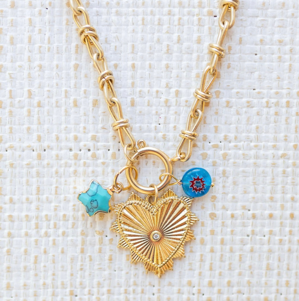 Picture of gold toggle chain with large gold heart pendant with a diamond in the center. Two charms are attached, one is a turquoise star and the other is a blue bead with a red ring in the center.