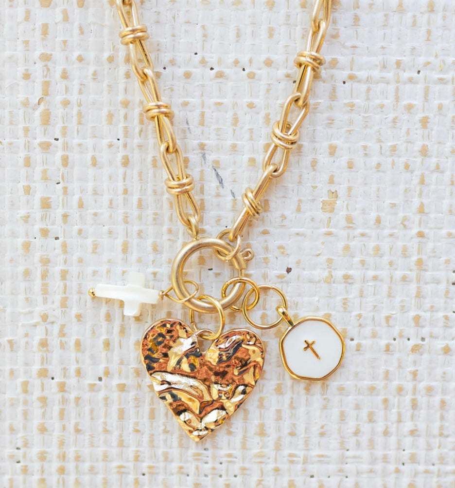 Picture of gold toggle chain with a textured gold heart pendant, a white cross charm and a white disc charm with a gold cross in the center.