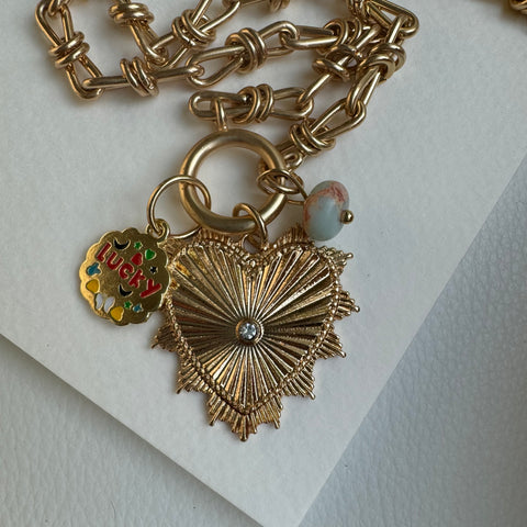 Picture of gold chain link charm necklace.  Necklace has a large gold heart pendant with a diamond at its center. Two smaller charms include a muted turquoise bead and a gold disc pendant with the word "Lucky" on it and small colorful inlays around it.